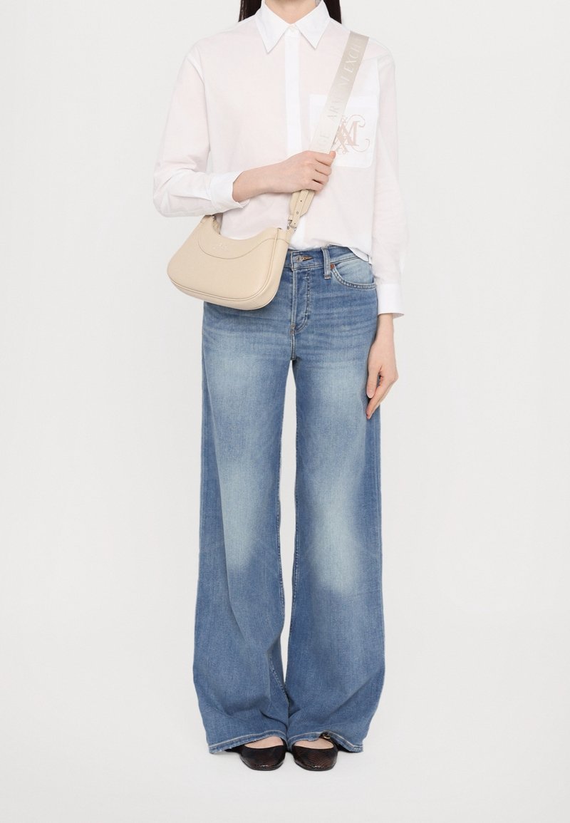 Borsa a tracolla in pelle beige con forma arrotondata, dotata di una tracolla robusta e un marchio discreto. Abbinata a una camicia bianca con bottoni e jeans a gamba larga.