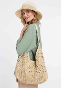 Beige háčkovaná kabelka přes rameno s otevřeným vzorem, širším dnem a pevným popruhem. Texturovaná tkanina v kombinaci se slámovým kloboukem.