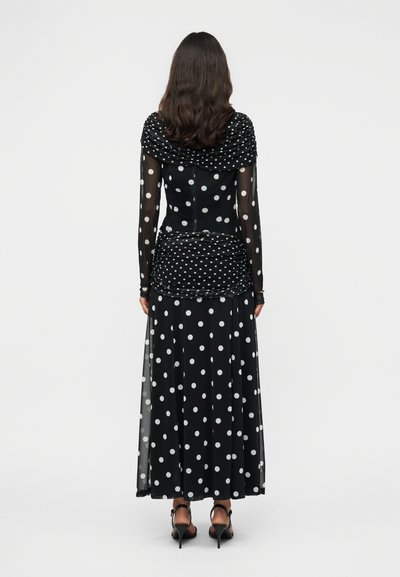 Μακρύ μαύρο φόρεμα με λευκές πουά, διαθέτοντας δομημένο πάνω μέρος με πιέτες και διαφανείς μανίκια, που καταλήγει σε φαρδιά φούστα.