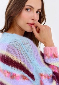 Wszechstronny sweter dziergany w wielu kolorach, charakteryzujący się luźnym, otwartym splotem. Pastelowe odcienie niebieskiego, różowego i brązowego tworzą poziome paski. Widoczna miękka faktura.