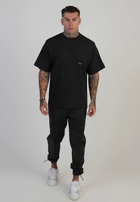 T-shirt noir à col rond avec manches courtes, associé à un pantalon de jogging noir fuselé. Présente un marquage discret sur la poitrine et des baskets blanches.