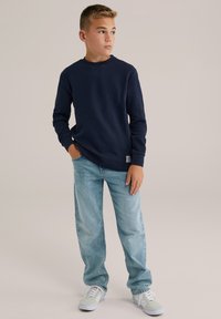 Tamsiai mėlynas ilgomis rankovėmis megztinis su tekstūruotu audiniu, derinamas su šviesiai mėlynomis džinsomis ir baltais sportbačiais, priekinis vaizdas.