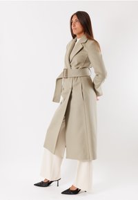 Cappotto lungo beige con vita cinturata, frontale doppiopetto e silhouette tagliata, abbinato a pantaloni a gamba larga crema e tacchi neri.