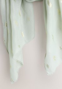 Tela verde claro con delicados motivos de hojas doradas y bordes deshilachados, colgando suelta sobre un fondo liso.