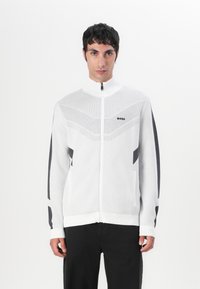 Bela zip-up jakna z unleženim vzorcem na zgornjem sprednjem delu in črnimi poudarki na rokavih ter žepih. Narejena iz mehkega pletenega materiala.