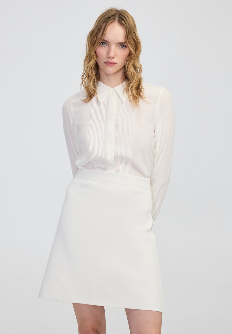 Weiße plissierte Bluse mit klassischem Kragen, kombiniert mit einem taillierten weißen Miniskirt. Glatte Texturen und schlichte Designdetails.