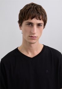 Camiseta negra de cuello en v hecha de algodón, con un sutil logo en el pecho izquierdo, mostrada sobre un fondo neutral.