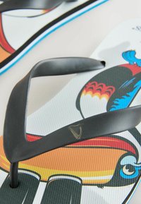 Tongs blanches avec des brides noires présentant des illustrations colorées d'oiseaux toucans et un petit logo doré de harpe sur la bride.