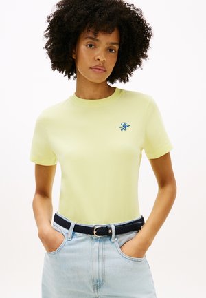 Kobieta o kręconych włosach, ubrana w jasnżółtą koszulkę z małym niebieskim logo oraz jasnoniebieskie dżinsy z czarnym paskiem.