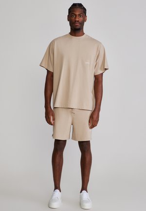 Hombre de pie mirando hacia adelante, vistiendo una camiseta beige de gran tamaño y pantalones cortos a juego, con zapatillas blancas, sobre un fondo liso.