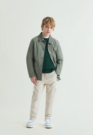 Niño joven con chaqueta verde, suéter verde oscuro, pantalones cargo beige y zapatillas blancas, de pie con una mano en el bolsillo.