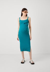 Robe de maternité en jersey teal avec des bretelles ajustables et une ceinture à nouer à la taille, présentée avec une longueur mi-mollet et une fente sur le côté. Associée à des sandales noires.