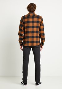 Bruin en zwart geruit flannel overhemd met een kraag met knopen, lange mouwen en een gebogen zoom, gecombineerd met zwarte slim-fit broek en sneakers.