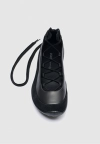Chaussure plate noire unique avec des brides élastiques croisées, mélange de cuir et de daim, et lacet prolongé non noué sur un fond clair.