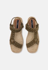 Paire de sandales plates avec semelles en corde tissée et lanières vert olive équipées d'œillets métalliques et de fermetures ajustables à la cheville.