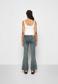Jeans de ganga azuis com flare, costura texturizada, bolsos traseiros e uma etiqueta na cintura, combinados com uma blusa de alças branca.