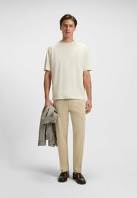 T-shirt crema a maniche corte abbinato a pantaloni beige e scarpe scure. Tiene in mano un giubbotto grigio a scacchi, posando su uno sfondo neutro.