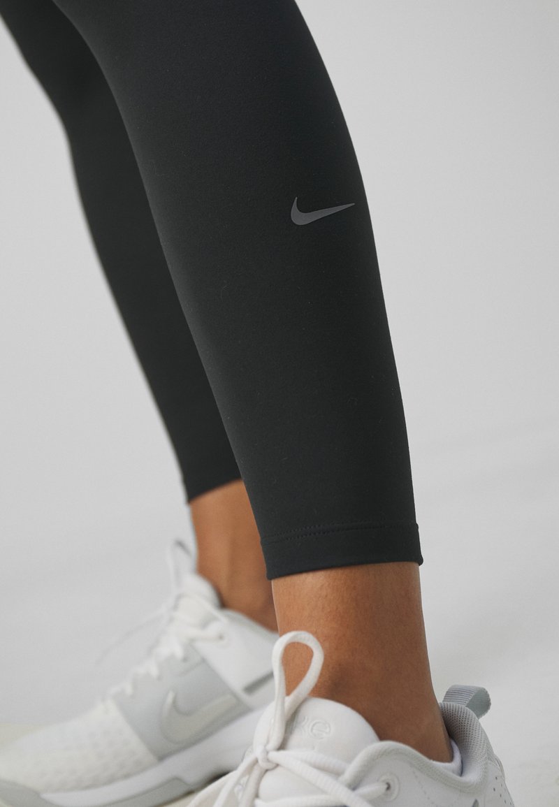 Leggings noirs avec un logo Nike réfléchissant, présentant une texture lisse et un design ajusté, associés à des chaussures de sport blanches.