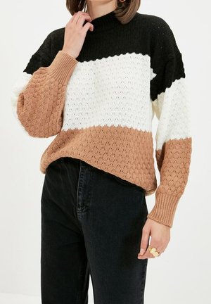 Osoba nosząca sweter w kolorowe bloki z czarnymi, białymi i beżowymi dzianymi sekcjami, w zestawieniu z czarnymi spodniami z wysokim stanem oraz złotymi pierścionkami na palcach.