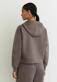 Sudadera con capucha marrón de corte holgado, tejido texturizado, dobladillo redondeado y mangas largas, que presenta un diseño minimalista y costuras sutiles.