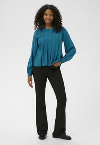 Teal blouse met geplooid ontwerp, lange mouwen en ronde halslijn, gecombineerd met zwarte flare pants en zwarte schoenen. Soepele textuur.