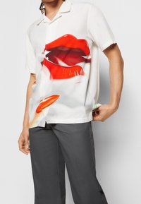 Lyhythihainen paita valkoisesta kankaasta, jossa on suuri grafiikka punaisista huulista ja oranssista huulipunan pullosta; napit edessä.