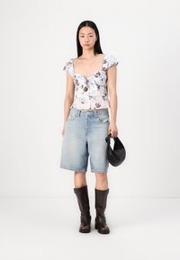Top corto floreale bianco con maniche corte, bermuda in denim azzurro chiaro e stivali al ginocchio in pelle marrone, mentre tiene una borsa nera.