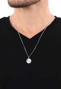 Silverfärgad halskedja med en rund berlock som har två fiskdesigns, fäst vid en tunn kedja. Bärs över en svart V-ringad t-shirt.
