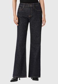 Donna che indossa jeans a vita alta, blu scuro, svasati con dettagli scintillanti sottili, abbinati a tacchi neri a punta e una maglietta nera.