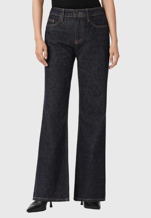 Donna che indossa jeans a vita alta, blu scuro, svasati con dettagli scintillanti sottili, abbinati a tacchi neri a punta e una maglietta nera.