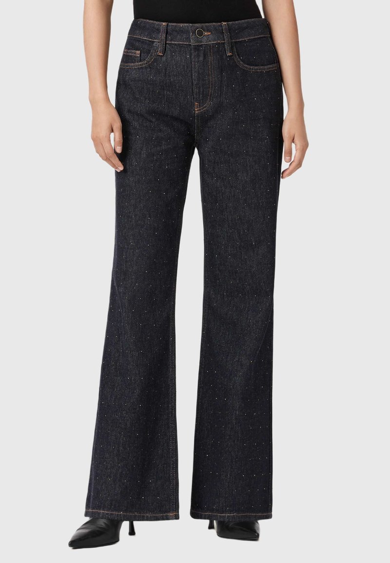 Donna che indossa jeans a vita alta, blu scuro, svasati con dettagli scintillanti sottili, abbinati a tacchi neri a punta e una maglietta nera.