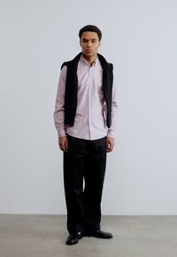 Άνδρας που στέκεται μπροστά σε μονόχρωμο τοίχο, φορώντας ανοιχτό ροζ πουκάμισο με κουμπιά, μαύρο παντελόνι, μαύρα παπούτσια και μαύρο πουλόβερ περασμένο στους ώμους.