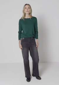 Kvinde med langt blondt hår iført en mørkegrøn langærmet skjorte, falmede sorte jeans og sorte støvler, står mod en ensfarvet baggrund.