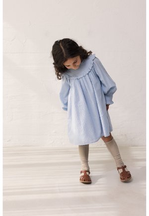 Dieťa s tmavými kučeravými vlasmi oblečené v svetlomodrých šatách, béžových podkolienkach a hnedých sandáloch stojace na svetlej podlahe pred jednoduchou stenou.