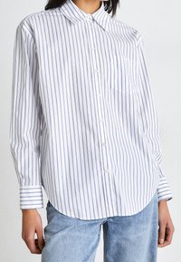 Košile s dlouhým rukávem na knoflíky v bílé barvě s modrými svislými pruhy, s límcem a přední kapsou, vyrobená z lehké bavlněné látky.
