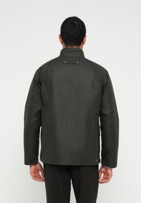 Σκούρο πράσινο μπουφάν με ψηλό γιακά, με ομαλή υφή και λεπτομέρειες με κουμπιά. Ελαφρώς φαρδιά εφαρμογή με τσέπες στα πλάγια.