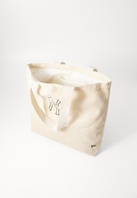Μπεζ βαμβακερή τσάντα ώμου με υφή κοτλέ, διαθέτοντας δύο υφασμάτινους ιμάντες και εκτυπωμένο λογότυπο στη μία πλευρά, επενδυμένη με απλό λευκό υλικό.