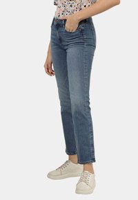 Calças de ganga azuis com corte reto, lavagem clara, design de cinco bolsos e textura subtil. Usadas com sapatilhas brancas.