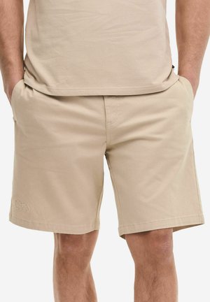 Hombre con pantalones cortos beige hasta la rodilla, con las manos en los bolsillos y camiseta beige a juego, de pie contra un fondo blanco.