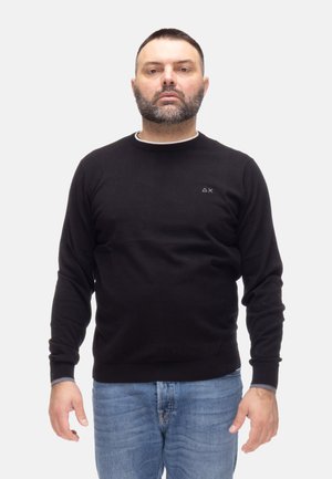 Uomo adulto con capelli corti e scuri e barba, che indossa un maglione nero e jeans blu, in piedi di fronte su sfondo bianco.