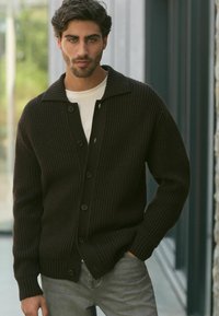 Cardigan en tricot marron foncé avec un col châle, doté d'une patte de boutonnage et d'une texture côtelée. Porté par-dessus une chemise blanche et un jeans gris.