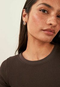 Brown ribbed shirt with a round neckline, worn by a model with medium-length dark hair and silver hoop earrings. Freckled skin is visible.