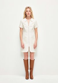 Vestido blanco de manga corta con cremallera frontal, cuello con volantes, dos bolsillos, tejido texturizado, combinado con botas de cuero marrón hasta la rodilla.
