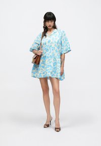 Ανοιχτό μπλε κοντό φόρεμα με μοτίβο φύλλων, φαρδιά εφαρμογή, V-λαιμόκοψη και κοντά μανίκια. Φορεμένο με καφέ τσάντα και ψηλοτάκουνα παπούτσια.