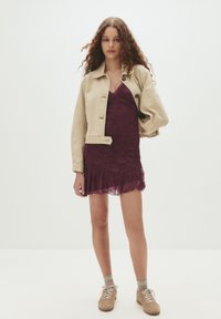 Frau mit langen lockigen Haaren, die eine beige Jacke über einem lila Spitzenkleid und beige Sneakers trägt, steht gegen einen schlichten hellen Hintergrund.