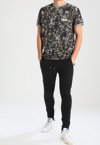 Kamouflage-mönstrad t-shirt i nyanser av grön och grå, med rund halsringning och "TRUE" logotyp. Slim-fit svarta joggers med ribbade accenter. Vita sneakers.