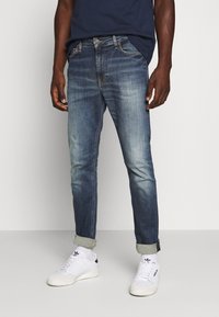 Blå denimjeans med smal passform, något blekta områden, uppvikta ärmar och flera fickor. Matchas med vita sneakers.