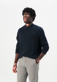 Navy knitted sweater with a textured pattern and a ribbed collar, paired with light gray tailored trousers featuring a subtle check pattern.