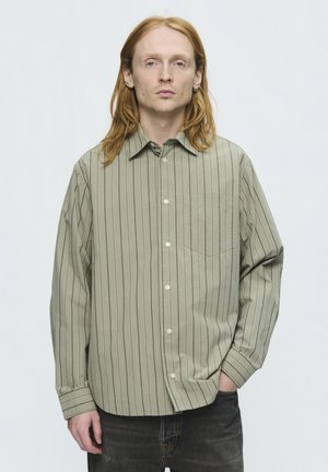 Jeune personne aux longs cheveux roux portant une chemise verte rayée boutonnée et un jean foncé, debout avec une main dans la poche.
