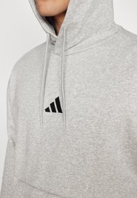 Sweatshirt cinza feito de tecido texturizado, com um bolso canguru, cordões e um logotipo triangular preto no peito.